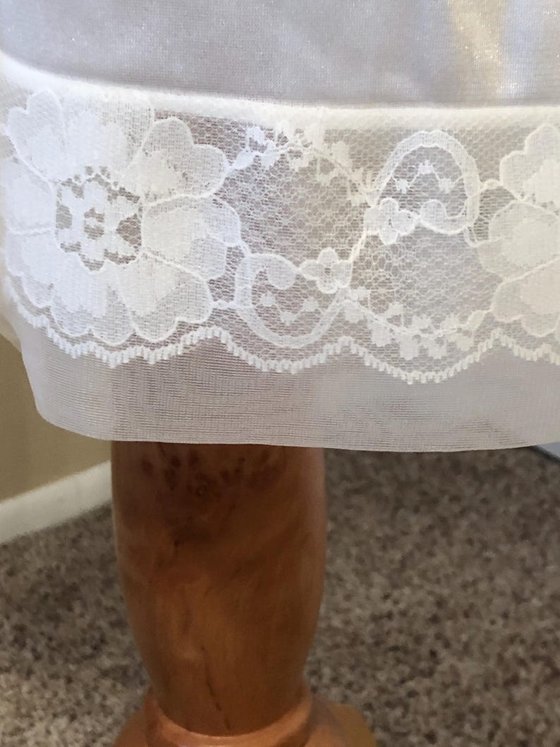 May include: A close-up view of a white slip, showcasing a detailed floral lace trim. The lace design includes repeating floral motifs and a scalloped border. The fabric is a soft, semi-transparent material.