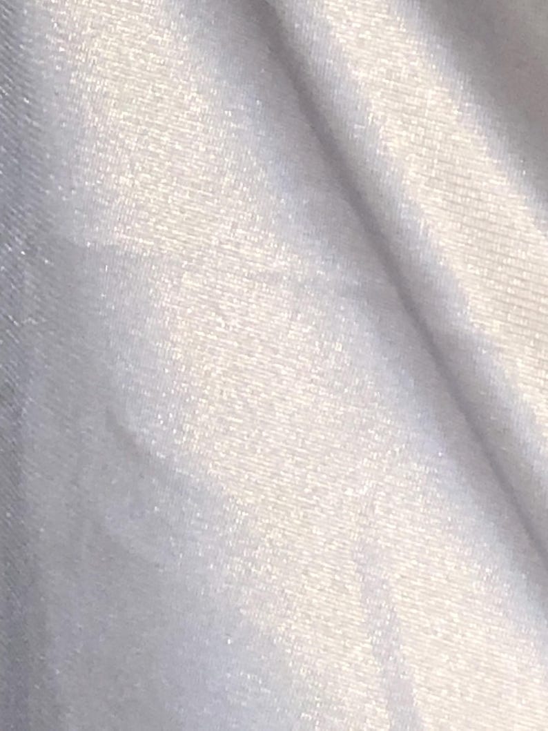 May include: Close-up of a white, textured fabric with a subtle sheen. The material appears to be soft and flowing, with visible diagonal folds and a slight shimmer, suggesting a luxurious textile.