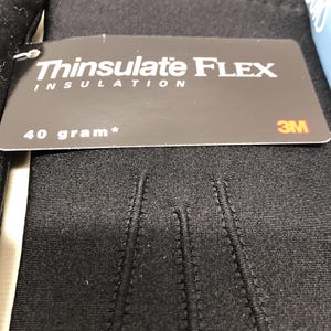 Op de afbeelding: Zwarte handschoenen met een label met de tekst "Thinsulate FLEX INSULATION" en "40 gram*". De handschoenen zijn gemaakt van een glad, donker materiaal en hebben zichtbare stiksels. Het 3M-logo is ook zichtbaar op het label.