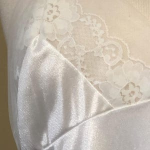 May include: A close-up of a white satin slip with delicate lace trim along the neckline. The fabric has a subtle sheen, and the lace features a floral pattern. The slip is displayed on a mannequin.