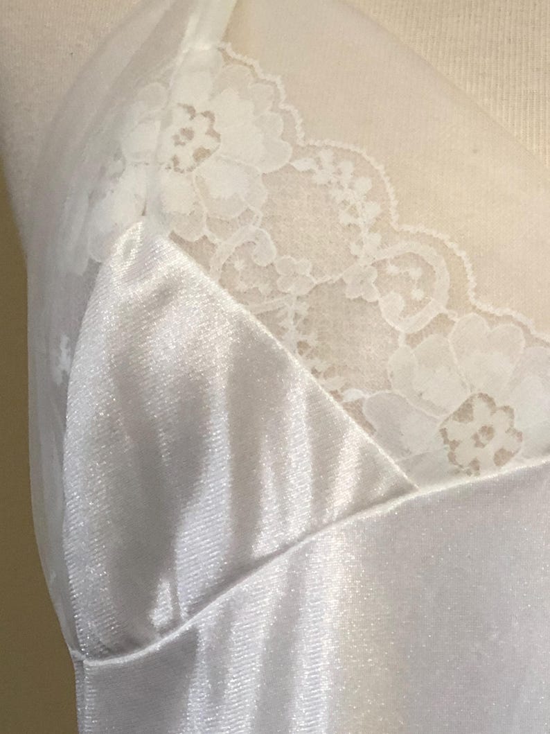May include: A close-up of a white satin slip with delicate lace trim along the neckline. The fabric has a subtle sheen, and the lace features a floral pattern. The slip is displayed on a mannequin.