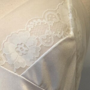 May include: Cream-coloured lingerie with floral lace detailing on the bodice. The fabric has a satin-like sheen. The design features a V-neckline and a soft, flowing silhouette. The overall aesthetic is elegant and delicate.