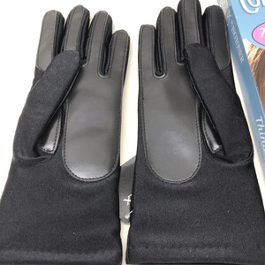 Op de afbeelding: Een paar zwarte handschoenen met leren accenten op de handpalmen en vingers. De handschoenen zijn gemaakt van een zachte, donkere stof en hebben een label. De handschoenen zijn ontworpen voor koud weer.