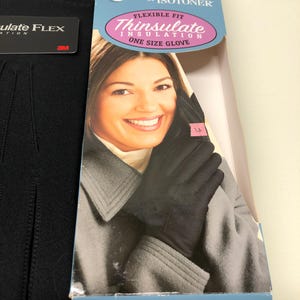Op de afbeelding: Zwarte Thinsulate ge&iuml;soleerde handschoenen met een flexibele pasvorm, one size. De verpakking toont "Contours by Isotoner" en "Thinsulate Insulation". De handschoenen worden getoond op een vrouw met een grijze jas.