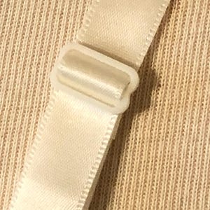 May include: Close-up of a cream-coloured bra strap with a white plastic adjuster. The strap is made of a smooth, satin-like material. The background is a textured, beige fabric.
