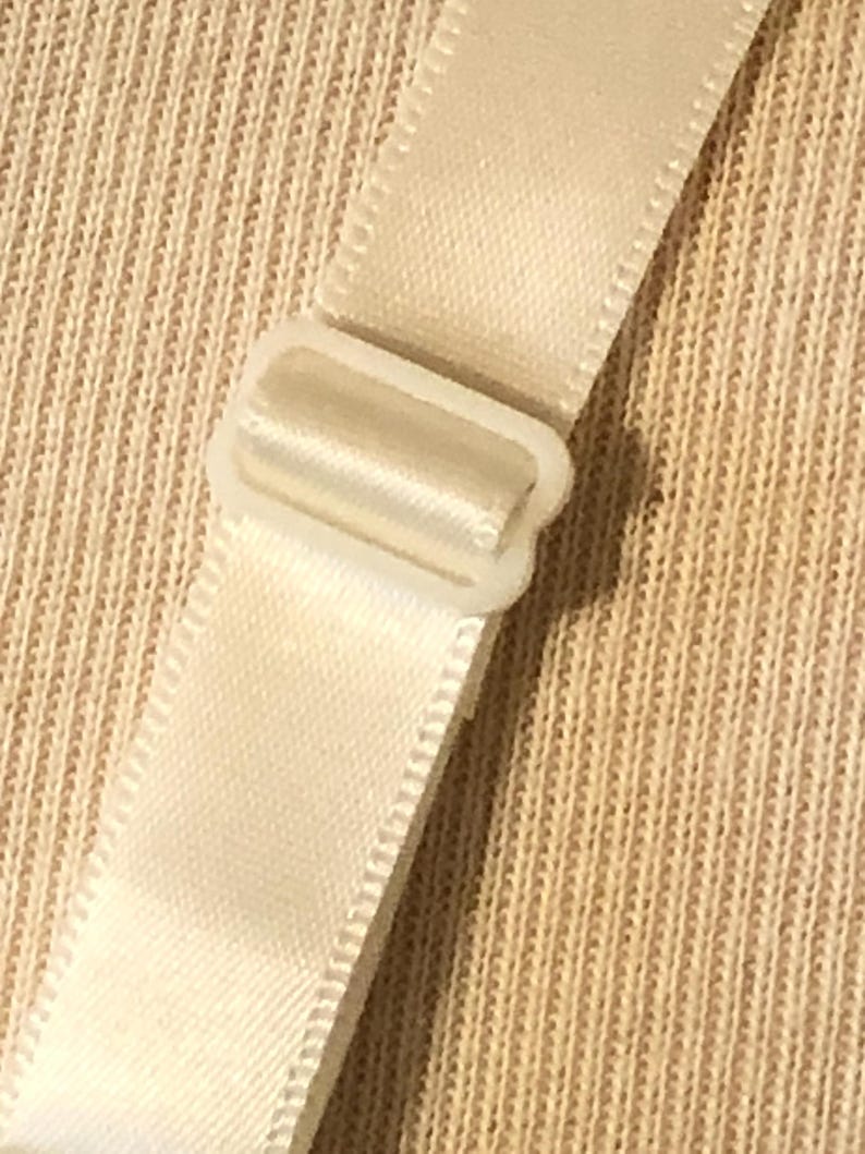 May include: Close-up of a cream-coloured bra strap with a white plastic adjuster. The strap is made of a smooth, satin-like material. The background is a textured, beige fabric.
