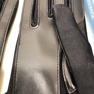 Op de afbeelding: Close-up van zwarte handschoenen met een combinatie van glad, glanzend materiaal en textuurstof. De handschoenen hebben een strak ontwerp met gestikte naden en verstevigde zones. De merknaam "ISOTONER" is gedeeltelijk zichtbaar op de achtergrond.