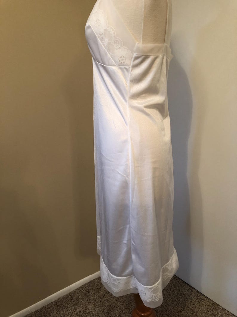 May include: A white satin slip dress with delicate lace trim along the neckline and hem. The dress is displayed on a mannequin, showcasing its flowing silhouette and vintage design. The slip features a V-neckline and a subtle sheen.