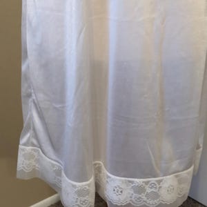 May include: A white slip dress with a delicate lace trim at the hem. The fabric appears to be a lightweight, flowing material. The lace trim features a floral pattern, adding a touch of elegance to the garment.