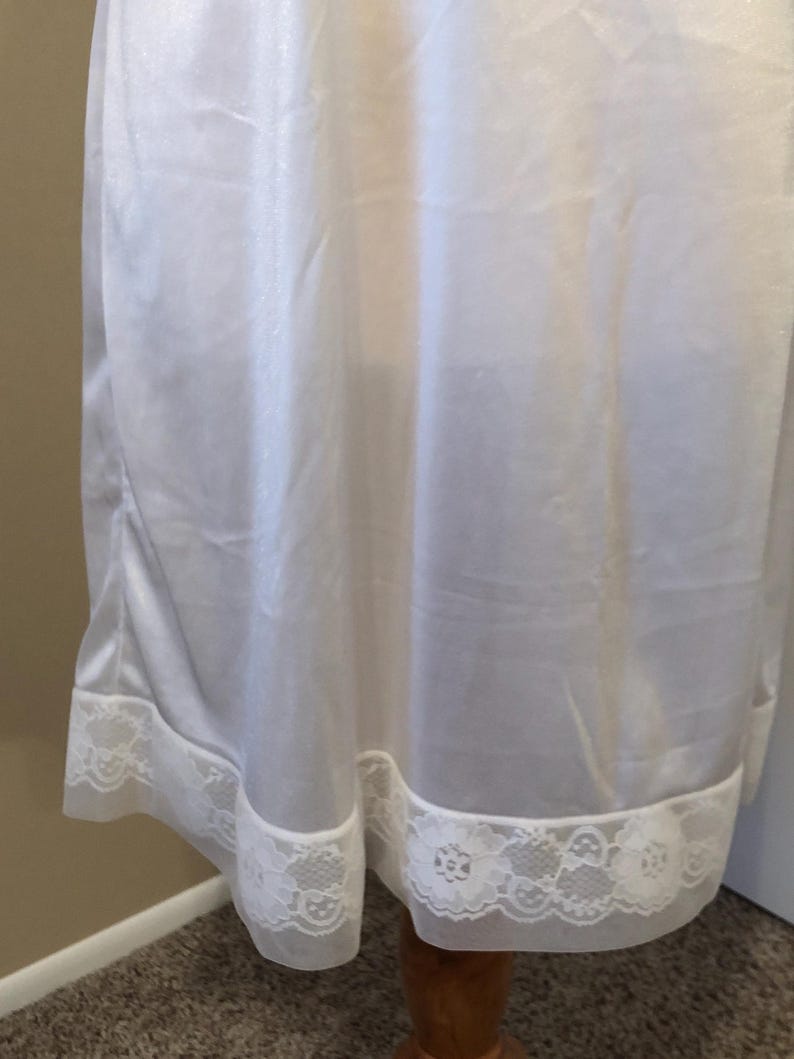 May include: A white slip dress with a delicate lace trim at the hem. The fabric appears to be a lightweight, flowing material. The lace trim features a floral pattern, adding a touch of elegance to the garment.