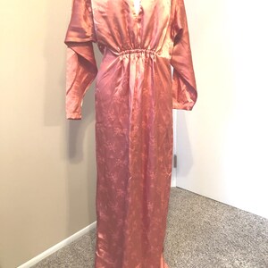 May include: A vintage, floor-length, peach-colored satin dress. The dress features a V-neckline, long sleeves, and an elastic waist. The fabric has a subtle floral pattern. The dress is a classic example of vintage fashion.