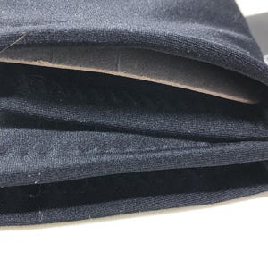 Op de afbeelding: Close-up van gevouwen zwarte stof, met lagen materiaal. De stof lijkt een zacht, mogelijk gebreid materiaal te zijn. De lagen zijn netjes opgestapeld, met een lichtgekleurde, mogelijk kartonnen laag zichtbaar tussen de stofvouwen.