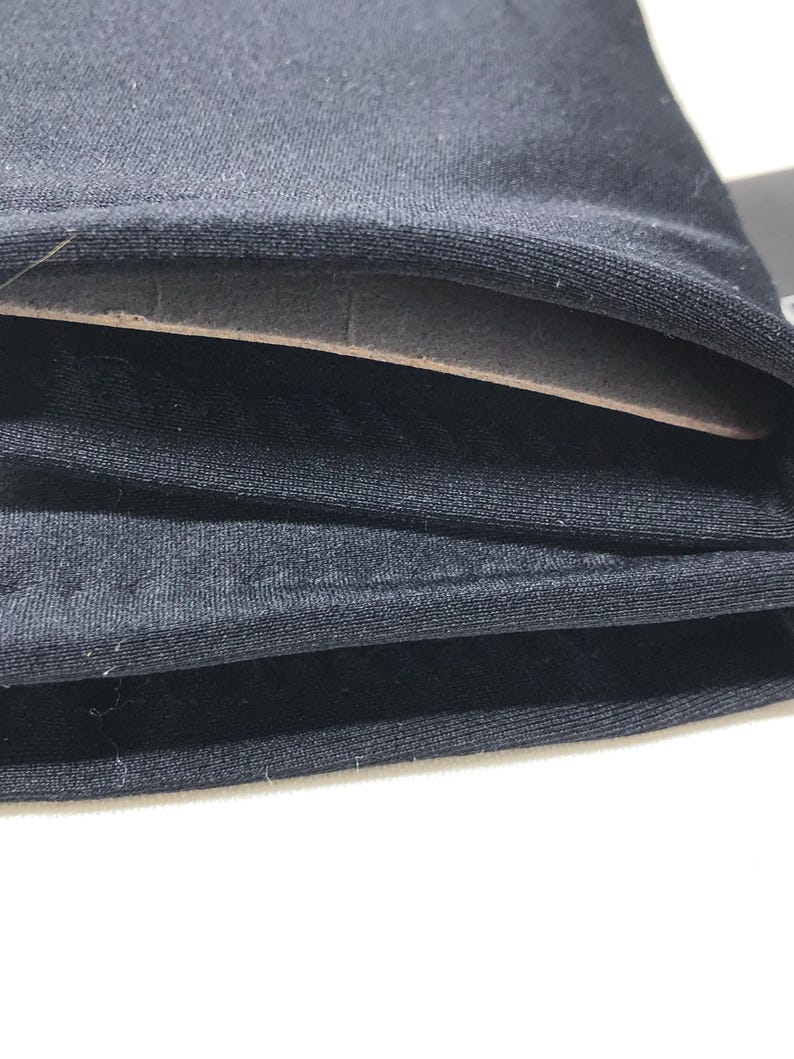 Op de afbeelding: Close-up van gevouwen zwarte stof, met lagen materiaal. De stof lijkt een zacht, mogelijk gebreid materiaal te zijn. De lagen zijn netjes opgestapeld, met een lichtgekleurde, mogelijk kartonnen laag zichtbaar tussen de stofvouwen.