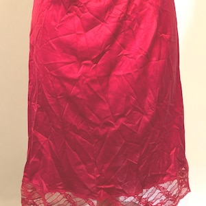 May include: A red satin slip skirt with a lace trim at the hem. The skirt is knee-length and has a smooth, shiny texture. The lace trim adds a delicate detail to the bottom of the skirt.