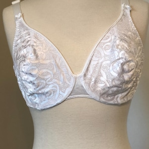 May include: White bra with a floral pattern. The bra is displayed on a mannequin. The bra has adjustable straps and a classic design. The fabric has a subtle sheen, and the overall look is delicate and feminine.
