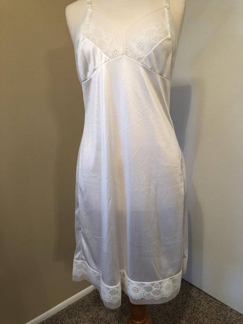 May include: A white satin slip dress with delicate lace trim on the neckline and hem. The slip has adjustable straps and a flattering shape. The dress is displayed on a mannequin.