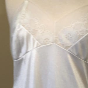 May include: A white satin slip with delicate lace detailing on the bust. The slip features adjustable straps and a V-neckline. The fabric has a subtle sheen, suggesting a smooth, comfortable texture. The overall design is simple and elegant.