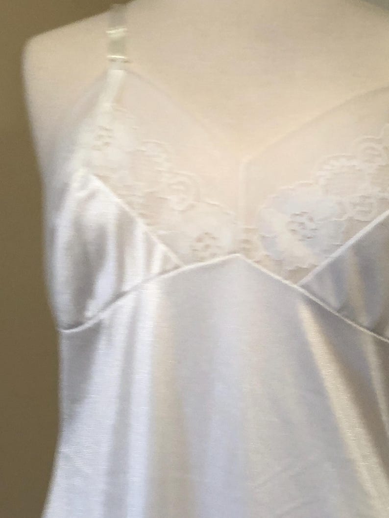 May include: A white satin slip with delicate lace detailing on the bust. The slip features adjustable straps and a V-neckline. The fabric has a subtle sheen, suggesting a smooth, comfortable texture. The overall design is simple and elegant.
