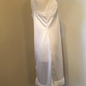 May include: A white satin slip dress with spaghetti straps and a lace trim along the neckline and hem. The dress is displayed on a mannequin. The slip dress is a classic lingerie piece.