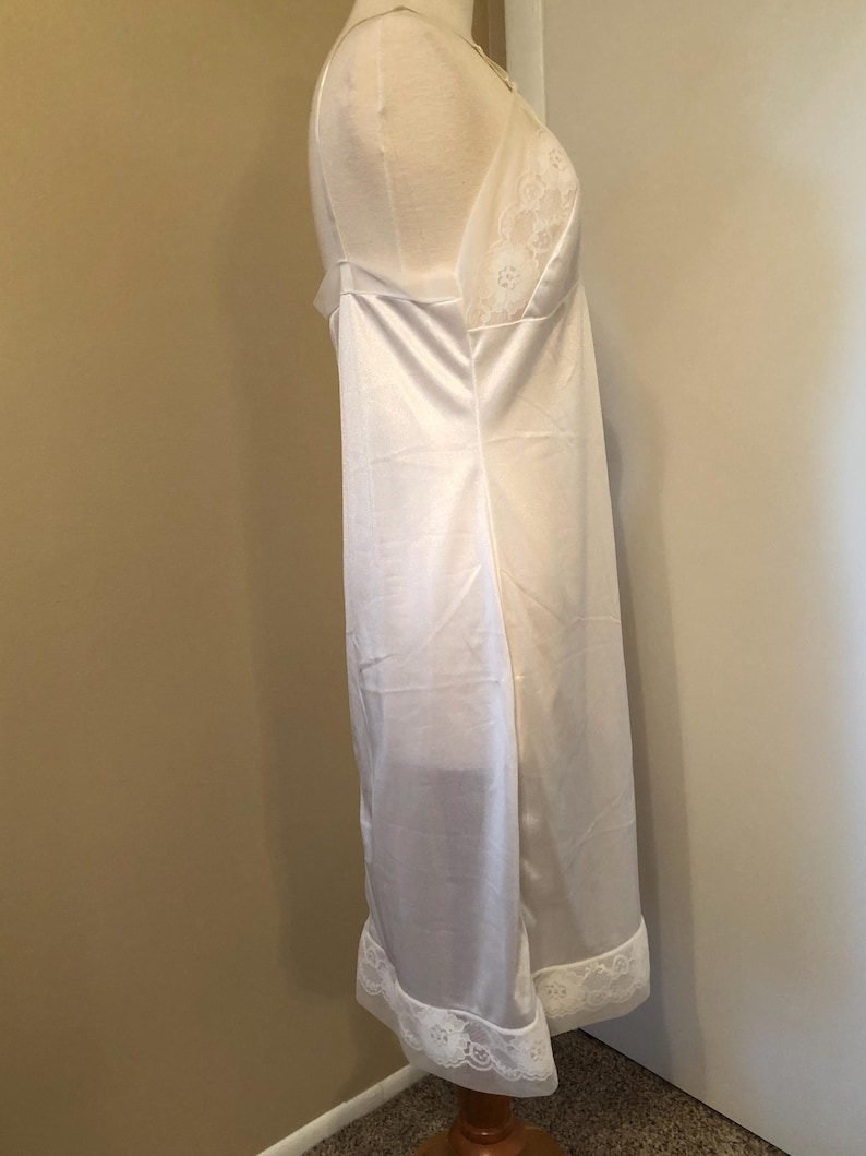 May include: A white satin slip dress with spaghetti straps and a lace trim along the neckline and hem. The dress is displayed on a mannequin. The slip dress is a classic lingerie piece.