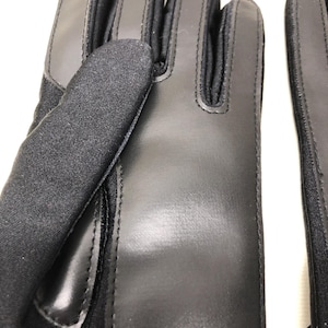 Op de afbeelding: Close-up van een paar zwarte handschoenen. De handschoenen zijn gemaakt van een combinatie van glad leer en een textuurstof. De stiksels zijn zichtbaar en de handschoenen lijken goed gemaakt.
