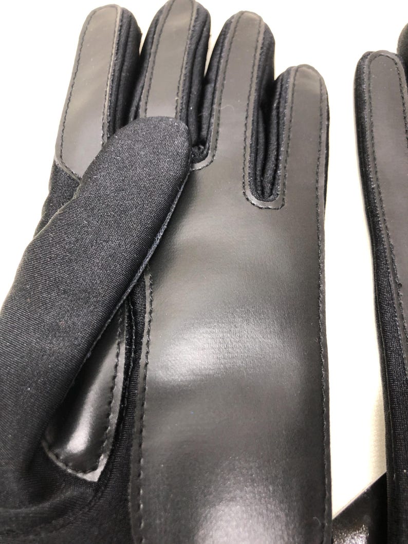 Op de afbeelding: Close-up van een paar zwarte handschoenen. De handschoenen zijn gemaakt van een combinatie van glad leer en een textuurstof. De stiksels zijn zichtbaar en de handschoenen lijken goed gemaakt.