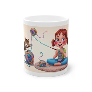 Könnte beinhalten: Eine weiße Keramiktasse mit einer bunten Illustration eines Mädchens, das mit Wollknäueln und einer Katze strickt. Das Mädchen trägt ein blaues Hemd und hat rote Haare. Die Katze ist braun und weiß.