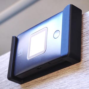Op de afbeelding: Een zwart rechthoekig apparaat met een scherm en een knop, gemonteerd op een lichtgekleurd houten oppervlak. Het apparaat wordt op zijn plaats gehouden door een zwarte beugel. Het scherm is verlicht en het apparaat lijkt een draagbare router te zijn.