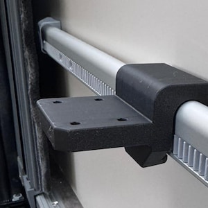 Könnte beinhalten: Silberfarbene Metallstange mit schwarzem Halter und grauer Stützstruktur. Die Stange hat ein geriffeltes Design und ist auf einer hellgrauen Oberfläche montiert. Die Halterung ist rechteckig mit mehreren Löchern. Die Stange ist ca. 50 cm lang.