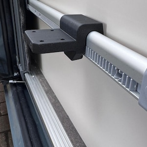 Puede incluir: Barra metálica plateada con un soporte de montaje negro fijado a una pared clara. La barra tiene un diseño estriado y forma parte de una estructura más grande, probablemente un componente de vehículo.