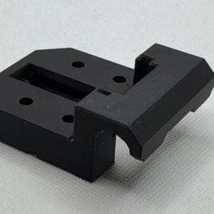 May include: Black plastic 3D printed part with a slot and several holes. The part has a curved section with a hook.