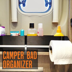 Könnte beinhalten: Ein schwarzes Regal mit verschiedenen Toilettenartikeln, darunter Flaschen und eine Tube, ist an einer Wand befestigt. Eine Rolle weißes Toilettenpapier befindet sich auf einem Spender. Der Text "CAMPER BAD ORGANIZER" ist orangefarben.