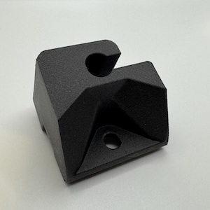 Puede incluir: Un soporte de esquina de plástico negro con dos agujeros. El soporte está diseñado para montarse en una pared u otra superficie.