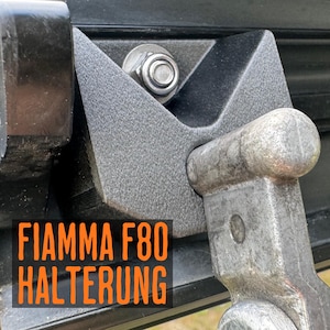 Könnte beinhalten: Eine schwarze, texturierte Halterung für eine Fiamma F80 Markise mit V-förmigem Design, befestigt mit einer Metallschraube und Mutter. Rechts befindet sich ein metallischer, verwittert wirkender Hebelarm, mit dem Text FIAMMA F80 HALTERUNG.