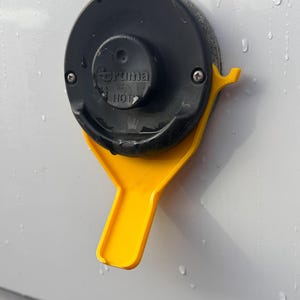 Może przedstawiać: Czarno-żółty zawór grzałki wody Truma z uchwytem. Zawór jest zamontowany na białej powierzchni.