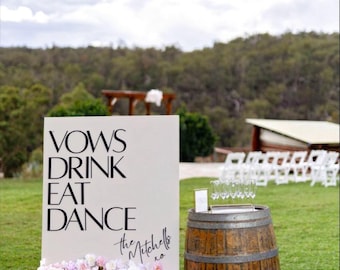May include: A rectangular off-white wedding sign featuring the black text VOWS DRINK EAT DANCE and the Mitchells xo signature. The base is decorated with a cluster of pink and white flowers. Beside it sits a wooden barrel and metal buckets.