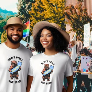May include: Two people wearing white t-shirts with the text "DON'T POKE THE BEAR" and a cartoon bear holding a map labeled "DEMOCRACY". The background includes a rainbow flag and a crowd of people.