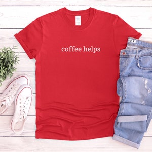 May include: Red t-shirt with the text "coffee helps" printed in white on the front. The shirt is laid flat on a white wooden surface with a pair of white sneakers and blue jeans.