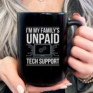 Könnte beinhalten: Schwarze Keramik-Tasse mit dem weißen Text "I'M MY FAMILY'S UNPAID TECH SUPPORT". Das Design beinhaltet eine Laptop-Grafik mit Zahnradsymbolen. Die Tasse hat einen schwarzen Henkel und wird von einer Person gehalten.