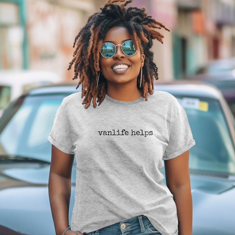 May include: A woman wearing a gray t-shirt with the text "vanlife helps" printed on it. She is wearing blue jeans and sunglasses.