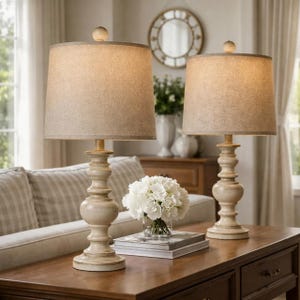 May include: Two beige table lamps with linen shades on a dark wood table. The lamps have a distressed, off-white finish and a classic, turned base design. A vase of white flowers and books are between the lamps. A sofa and mirror are in the background.