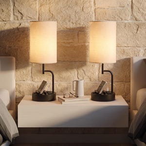 May include: Two black table lamps with cylindrical beige fabric shades and integrated charging bases. Each lamp features a black metal arm, a phone stand slot, and a USB port. The lamps are positioned on a white floating shelf between two beds.