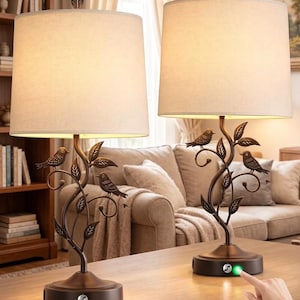 May include: Two bronze table lamps with cream shades. Each lamp has a decorative branch design with small bird figurines. The base of each lamp has a touch-sensitive power button. The lamps are on a wooden table.