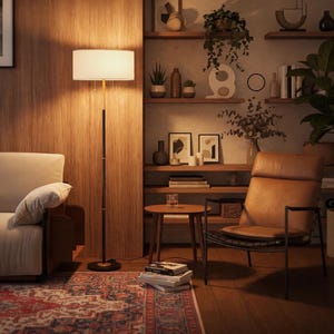 May include: A tall floor lamp with a cylindrical white fabric shade, a thin black metal pole, and a circular base. The lamp emits a warm glow, illuminating a living room space featuring a sofa, a leather armchair, and wooden shelving.