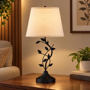 May include: A table lamp featuring a black metal base with a curved stem adorned with metal leaves. The lamp is topped with a tapered, off-white fabric drum shade. The base includes a small power switch located near the bottom of the stand.