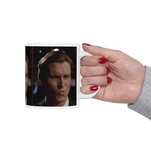 Puede incluir: Taza de cerámica blanca con la imagen de un hombre con expresión severa. La taza es sostenida por una mano con esmalte de uñas rojo. La taza es de tamaño estándar para café o té.