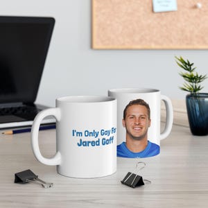 May include: Two white ceramic mugs. One has the text "I'm Only Gay For Jared Goff" in blue. The other mug features a colour portrait of a smiling man in a blue jersey. The mugs are on a light surface.