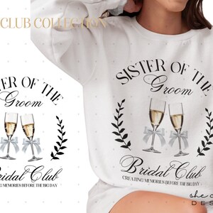 Puede incluir: Sudadera blanca con texto negro que dice "Hermana del novio Bridal Club Creating Memories Before the Big Day". El texto está rodeado de diseños florales negros y dos copas de champán con lazos plateados.