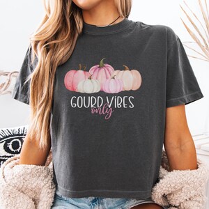 Puede incluir: Camiseta gris oscuro con un diseño de acuarela de calabazas rosas y blancas. El texto "GOURD VIBES only" está impreso debajo. La camiseta tiene mangas cortas y cuello redondo.