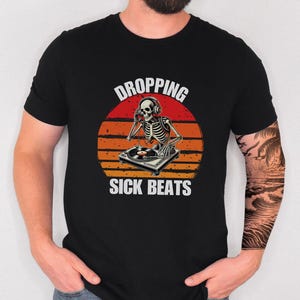 May include: Black t-shirt with a skeleton DJ graphic wearing headphones and playing a record. The design includes the text "DROPPING SICK BEATS" against a sunset background.